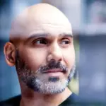 A bald man with a beard looking through a window with a thoughtful expression.